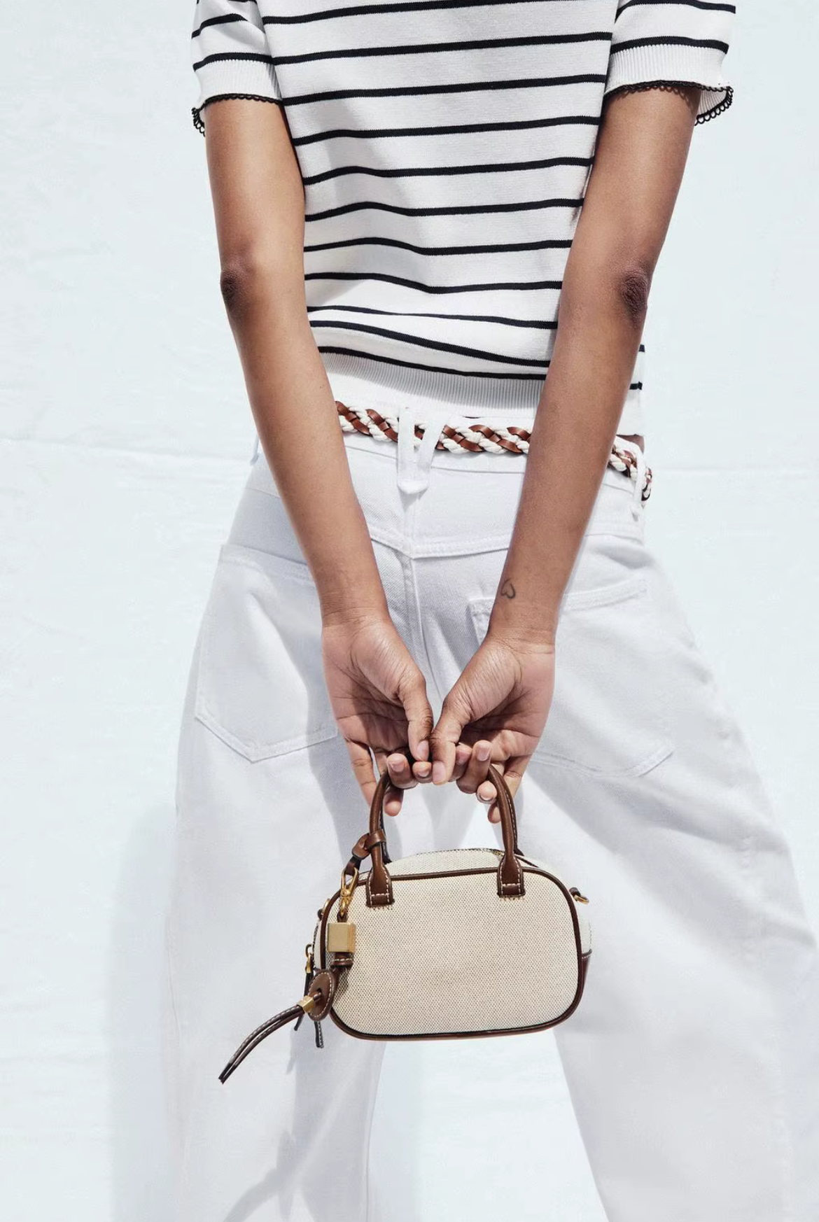 Person holding a beige handbag wearing a striped shirt and white pants on a light background