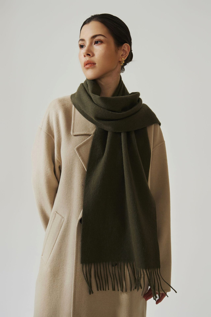 Woman wearing a beige coat and green scarf with fringe on a plain background