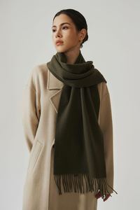 Woman wearing a beige coat and green scarf with fringe on a plain background