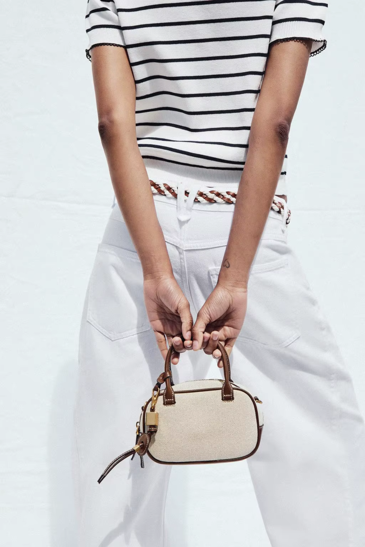 Person holding a beige handbag wearing a striped shirt and white pants on a light background