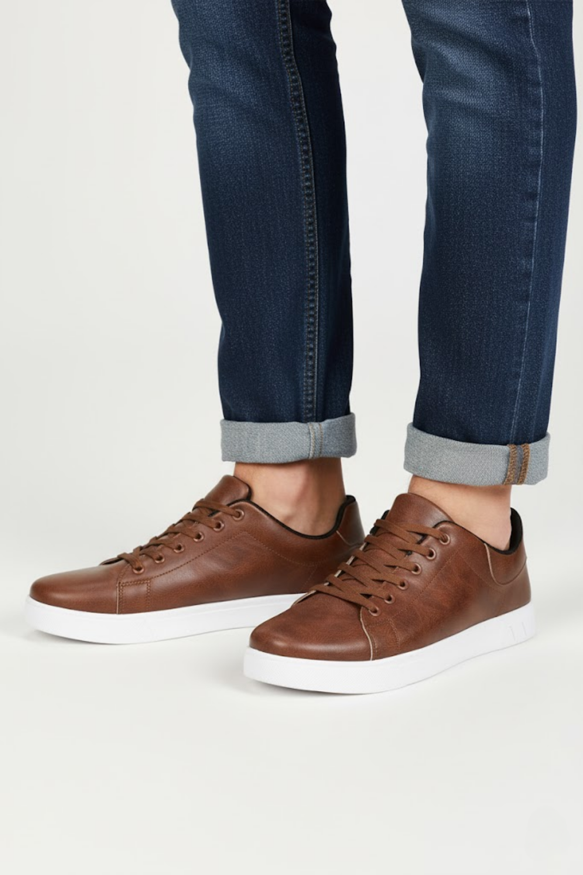 Brown leather sneakers worn with rolled-up jeans on a white background