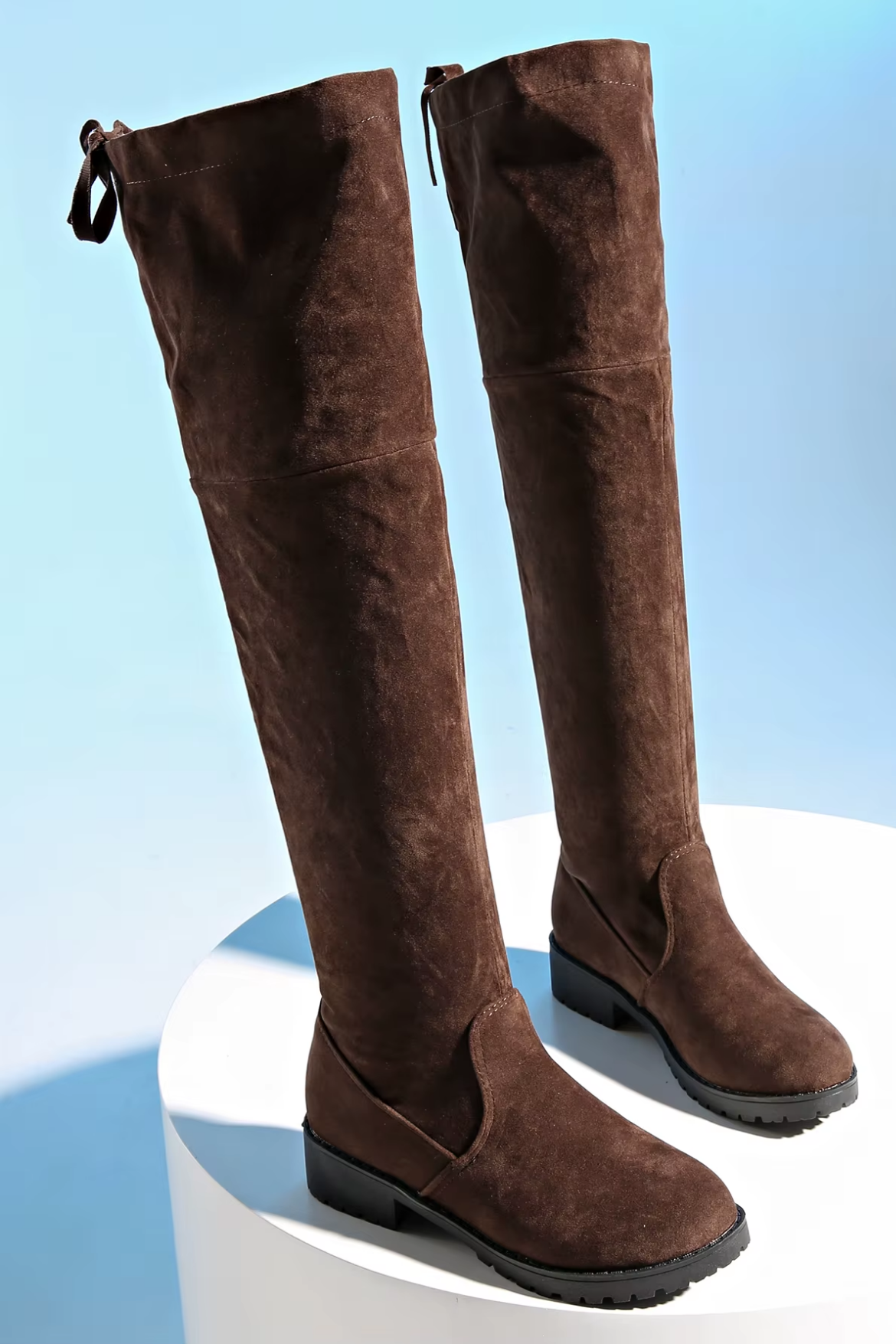 Brown suede knee-high boots on a white platform with a light blue background