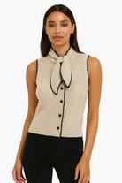Woman wearing a beige sleeveless knitted top with black buttons and trim on a white background