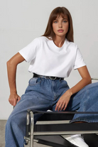 Woman sitting on a chair wearing a white t-shirt and blue jeans against a plain background