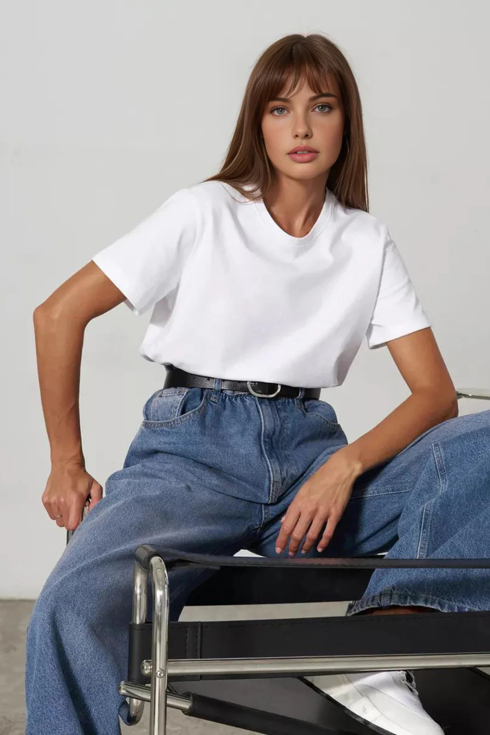 Woman sitting on a chair wearing a white t-shirt and blue jeans against a plain background
